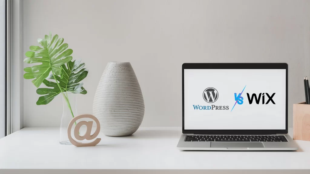 Laptop-on-a-table-displaying-WordPress-vs-Wix-text-with-a-vase-plant-and-decorative-@-symbol-nearby